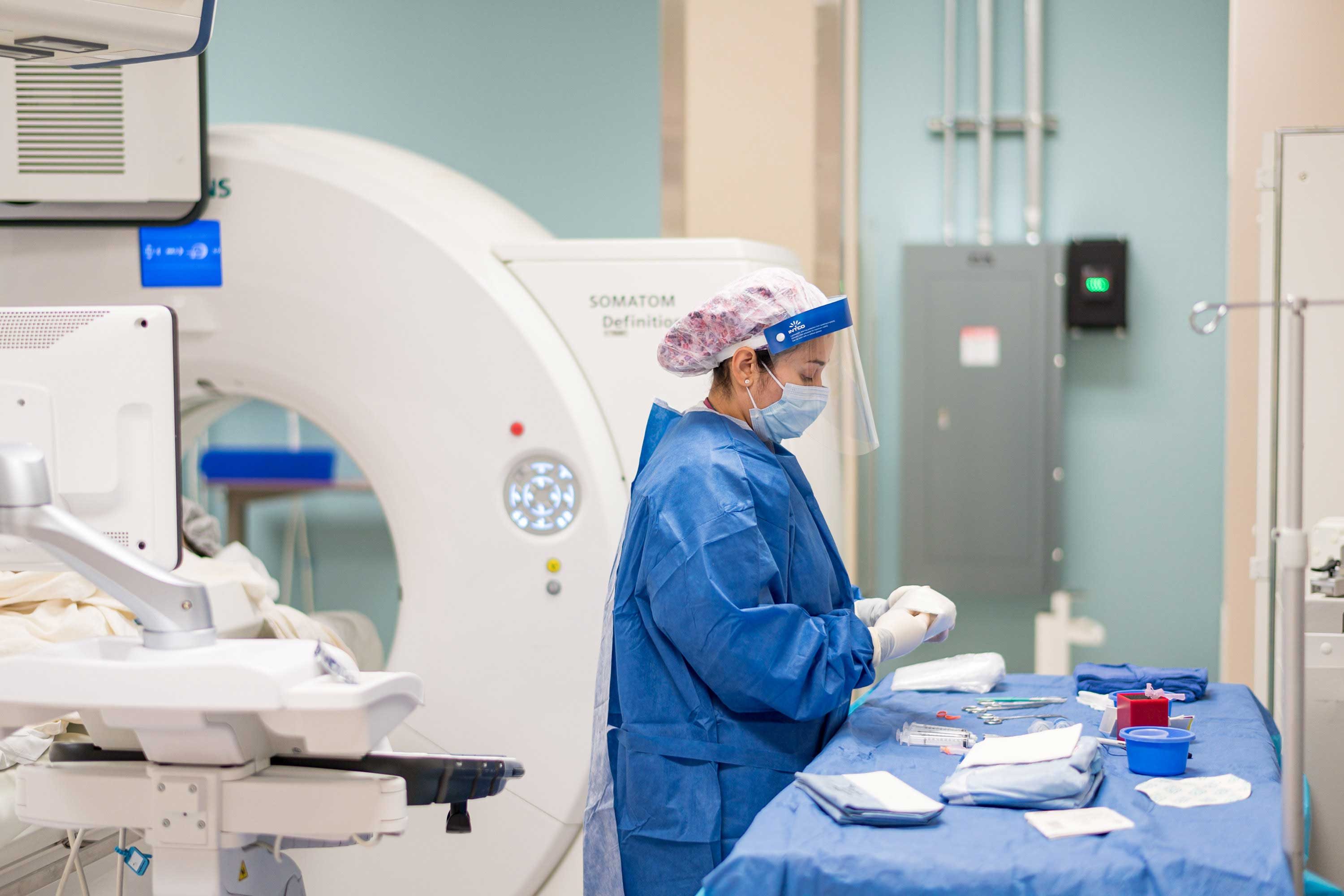 A person dressed in full scrubs and PPE stands in front a table preparing utensils for an interventional radiology procedure. Behind her is a scanning machine.