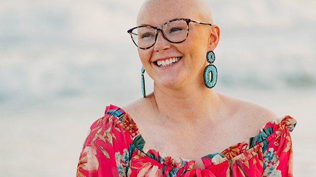 Stage IV Hodgkin lymphoma survivor Mallory Parrish, wearing glasses and a red floral top and big turquoise earrings, smiles while looking off in the distance.