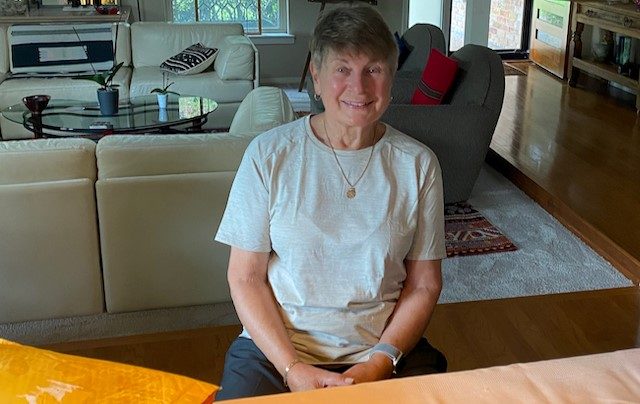 Andrea Spica smiling sitting in a chair. A beige sofa and glass coffee table with plants on it sit behind her.
