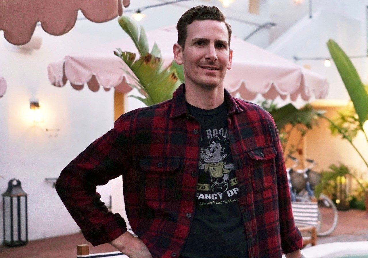Esophageal cancer survivor Ross Bernkrant stands on an outdoor patient with his hand on a table and a pink table umbrella hanging overhead. He is wearing a black t-shirt and black pants with a red and black flannel button shirt over it, unbottoned.