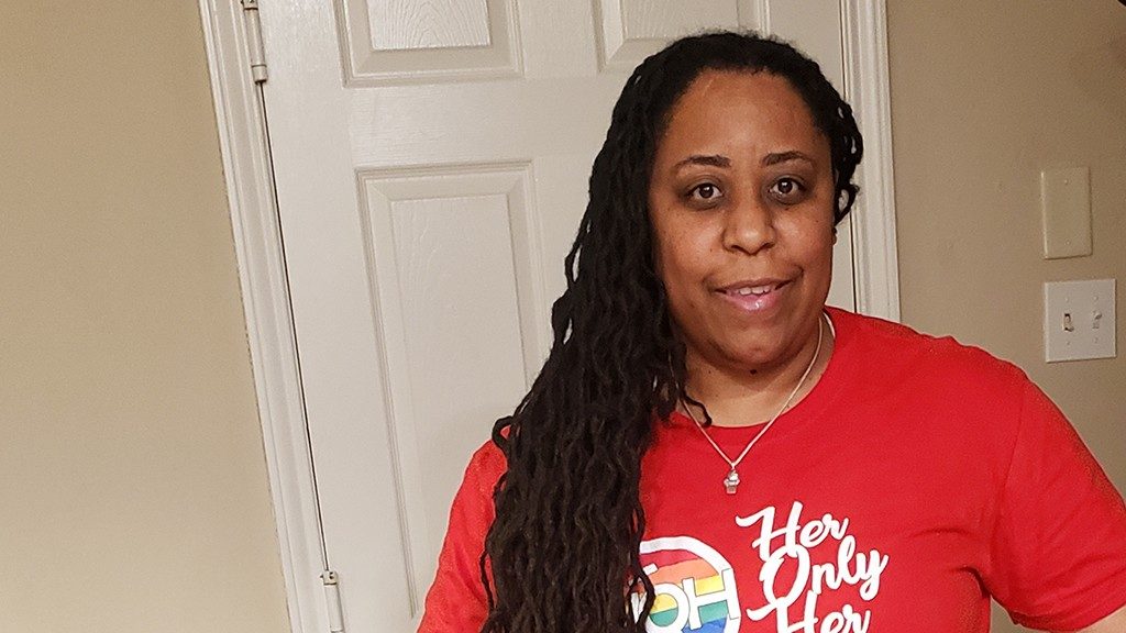 Breast cancer survivor MaKieba Hatton wears a red shirt and stands in front of a white door.