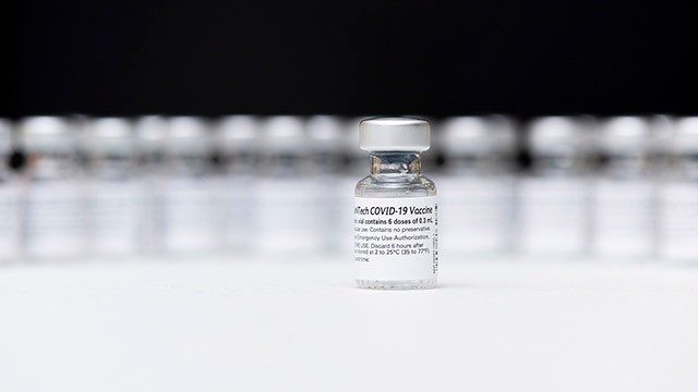 Clear vials of COVID-19 vaccine lined up against a black background, with one vial sitting in front of the others in the middle