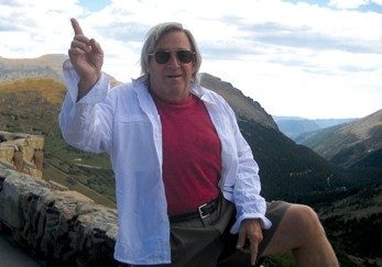 Gerry McKim pointing to the sky and standing outside a scenic mountain top.