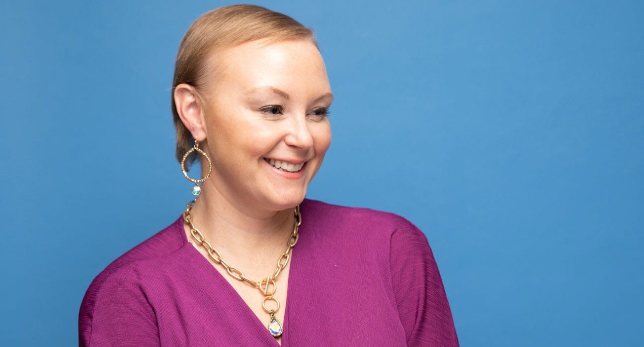 Hodgkin lymphoma survivor Mallory Parrish smiles while wearing a pink blouse, gold chain necklace and large earrings, while standing in front of a blue background.