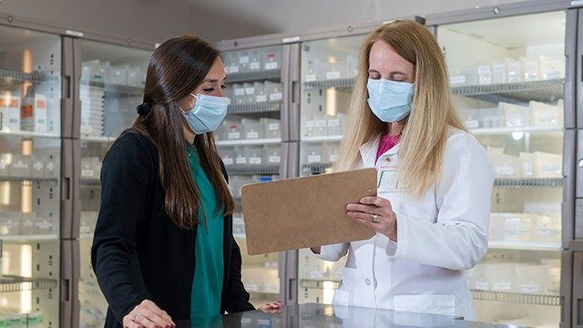 Pharmacy Manager Stefani Gautreaux (right) holds up a clipboard while speaking with pharmacy resident Lauren Goldberg in the pharmacy. Both women have long hair and are wearing medical-grade masks.
