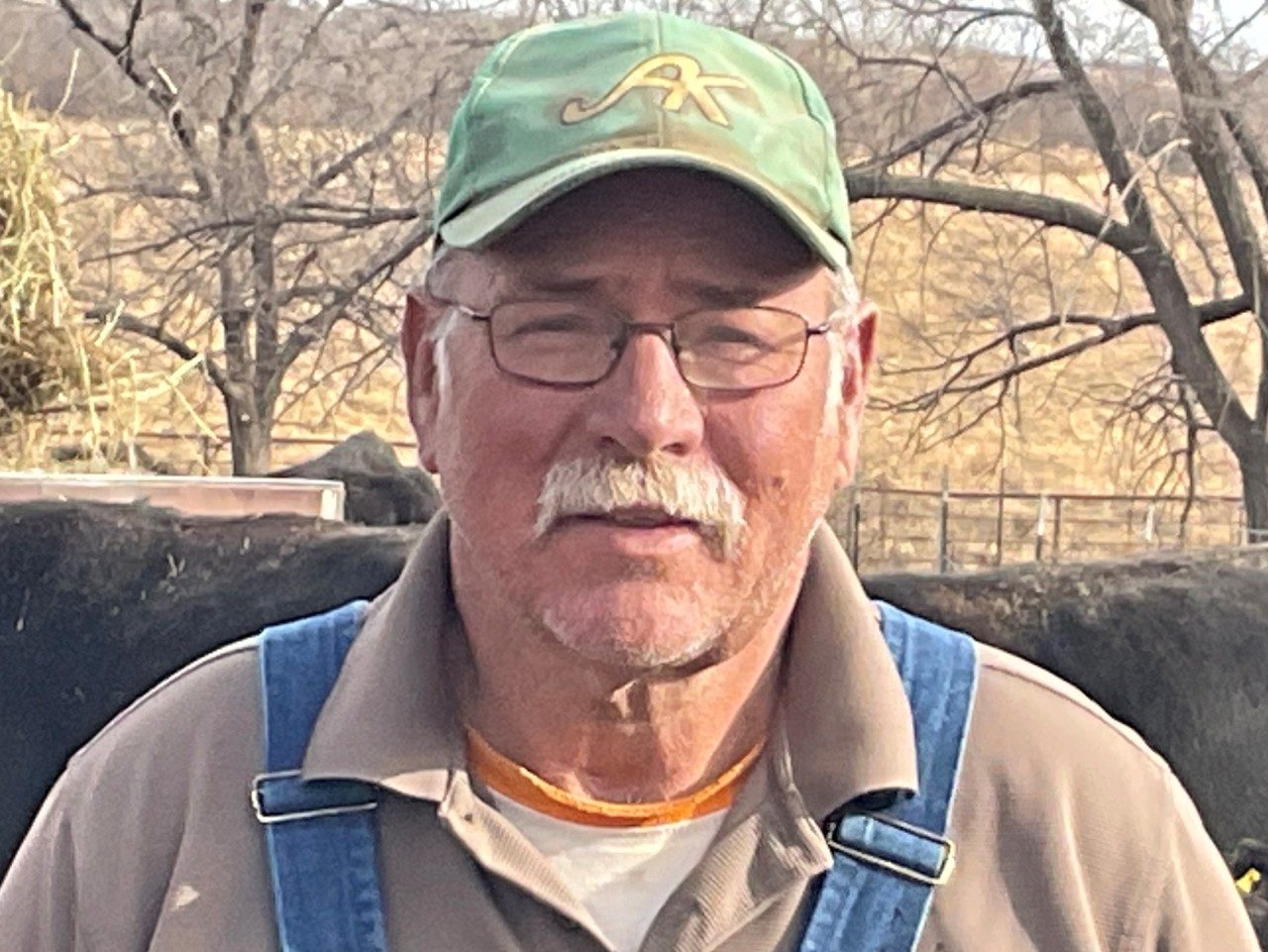 Dane Clark, wearing a baseball cap and overalls, stands outside in a pasture with cows behind him