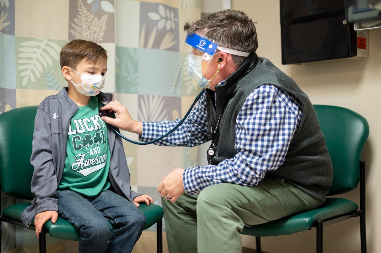 Medical oncologist Douglas Harrison, M.D., listens to the heart of pediatric rhabdomyosarcoma patient Sawyer Hack, sitting down in a clinic examination room.