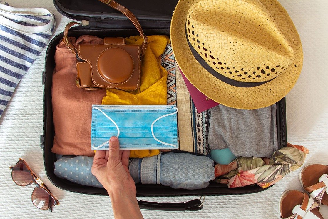 Bird's eye view of open suitcase packed for vacation with hand placing face mask inside