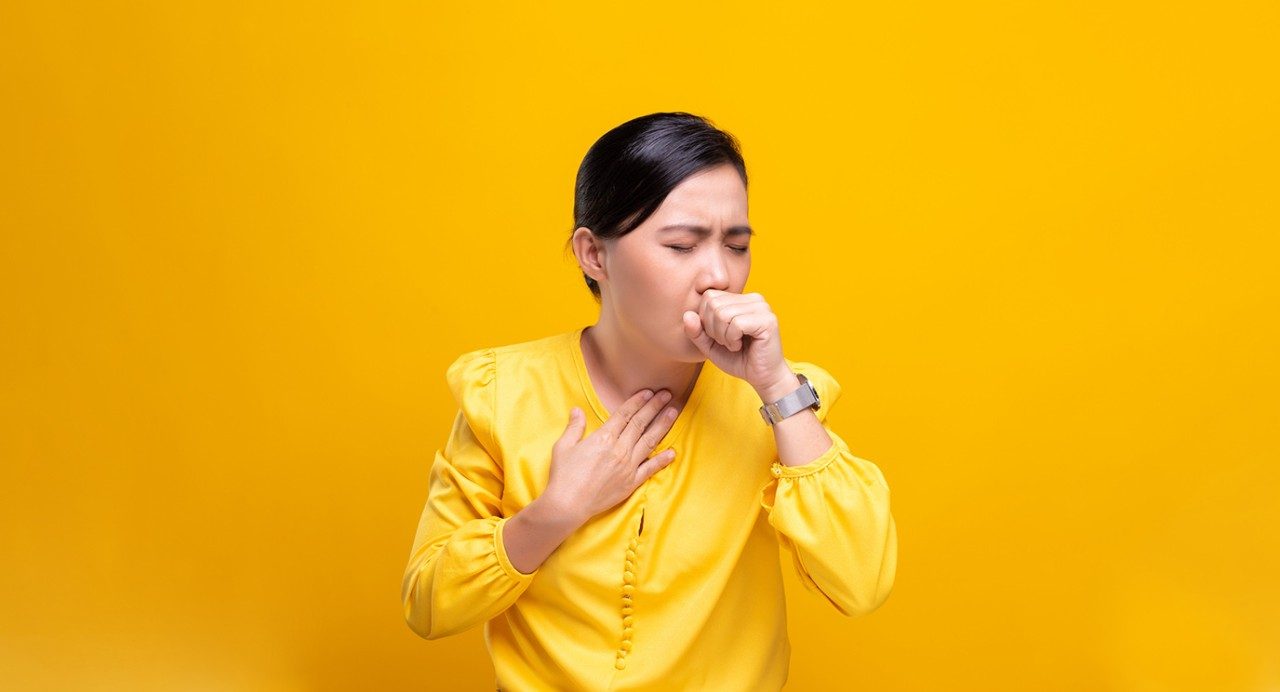 Person in bright yellow long-sleeve blouse coughing against a deep yellow background 