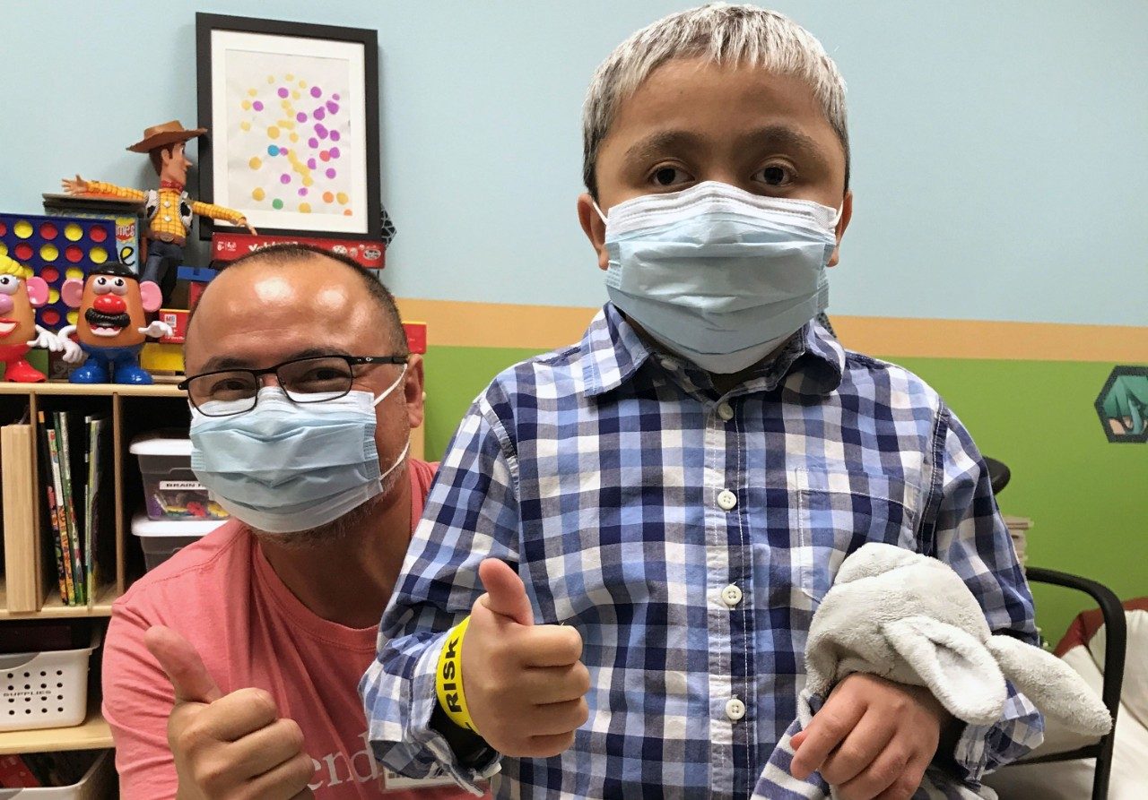 Pediatric brain tumor survivor Ben Gadot gives a thumbs up while standing in front of child life specialist Leon Benavides at MD Anderson Proton Therapy Center. 