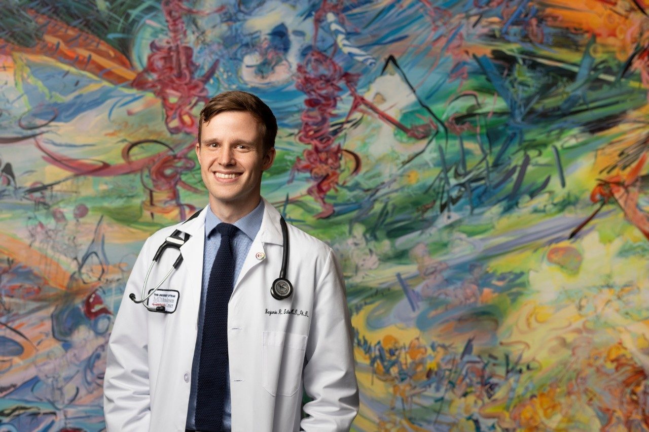 Benjamin Schrank, M.D., Ph.D., stands in front of a painting with his white coat on.