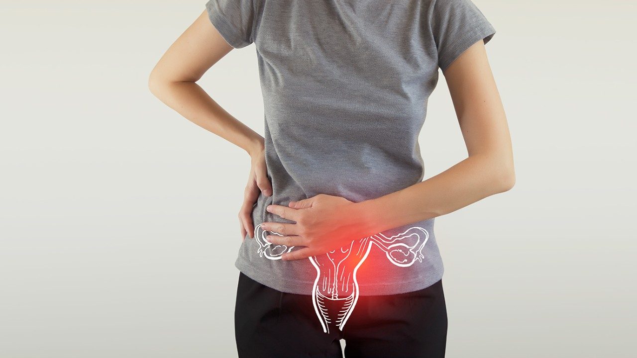 Neck to hips photo of slender, light-skinned woman in gray T-shirt and black pants, holding her abdomen with one hand and her hip with the other, over anatomical drawing of female reproductive system with red glow indicating pelvic pain