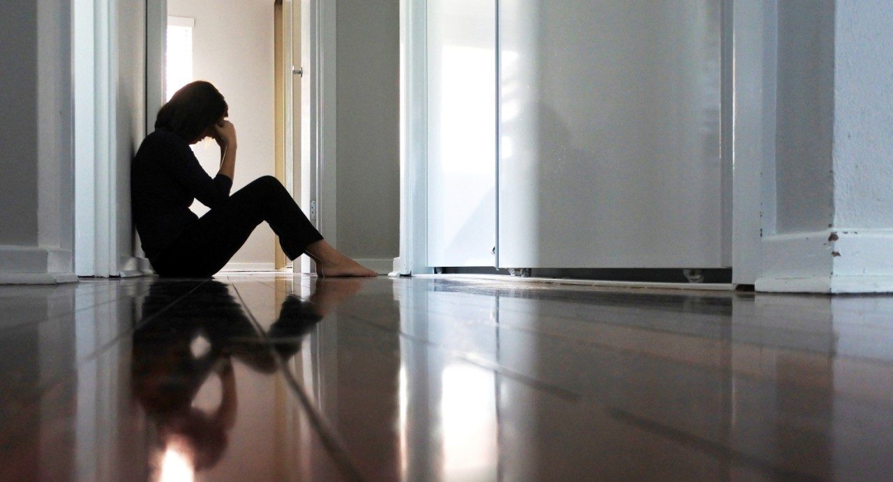 Person leans against wall with head in hands while sitting on shiny wood floor in empty home