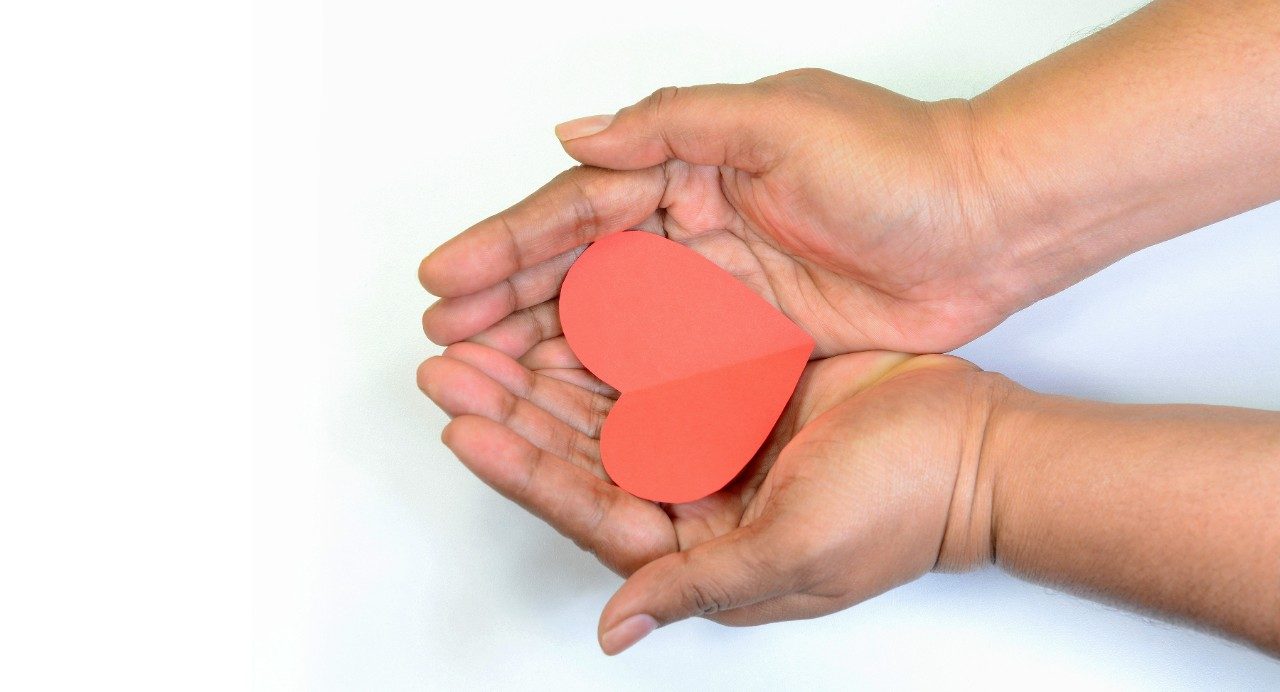 Close-up of hands holding red paper heart together in their upturned palms against a white background