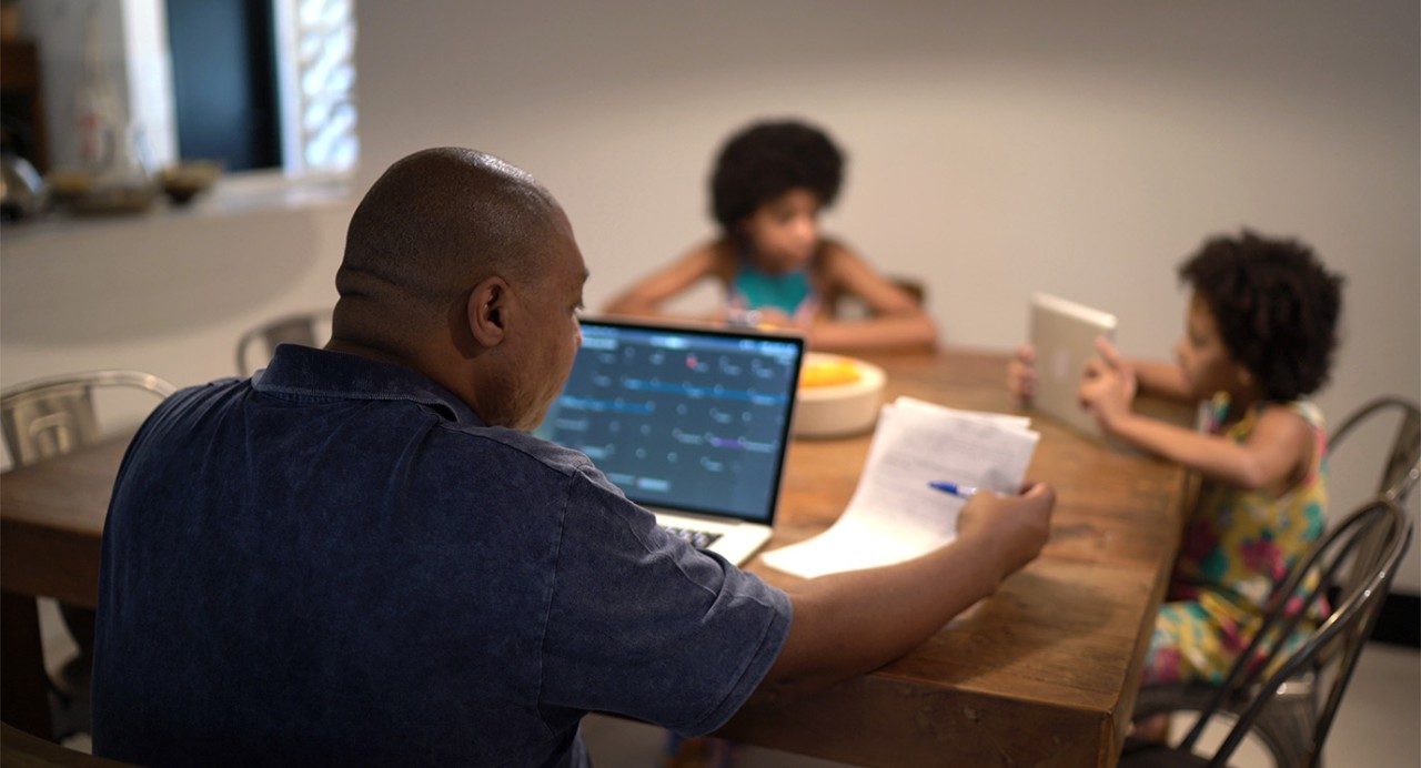 A middle-aged man sits at the table looking at his computer while two kids sit across from him