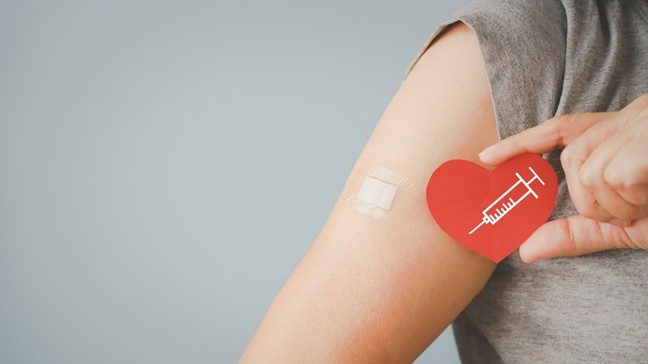 A person wearing a grey t-shirt with the sleeve pushed up to show a clear bandage on their arm. In the other hand, they hold a red heart-shaped sticker with a white graphic of a vaccine.