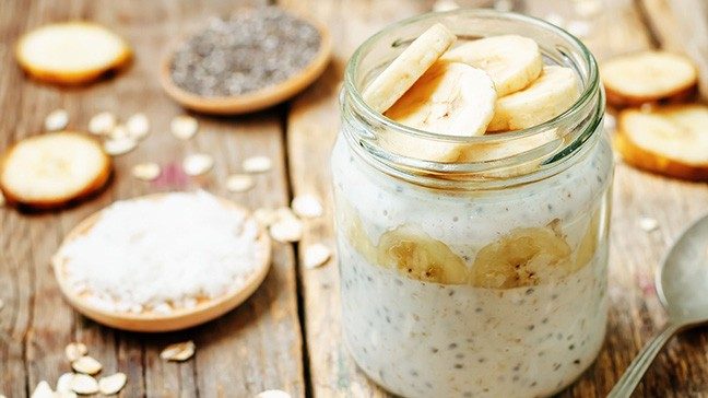 Glass jar with chia seeds, sliced banana and oats on a wooden table alongside a spoon and small dishes of chia seeds and other toppings.