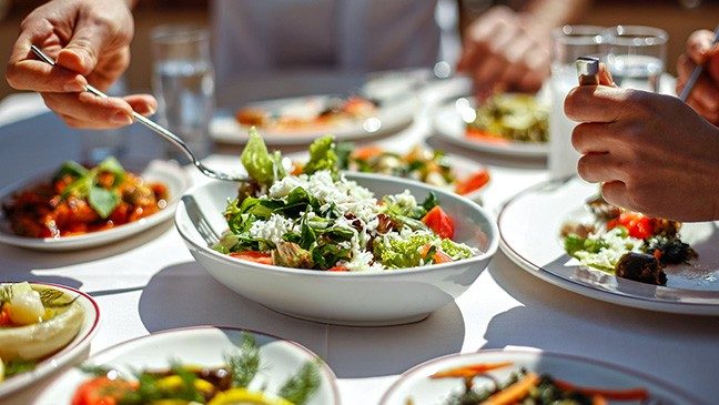 Two sets of hands hold silverware over white dishes containing vegetables on a white table