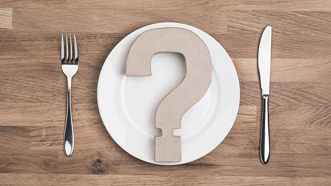 A white plate with a cardboard question mark placed on it sits on a wooden surface. A silver fork and knife sit on either side of the plate.