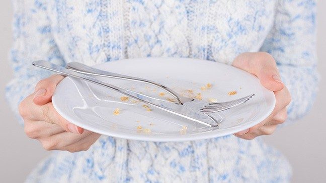 Person in blue and white cable knit sweater holds an empty white plate holding crumbs and a silver fork and knife