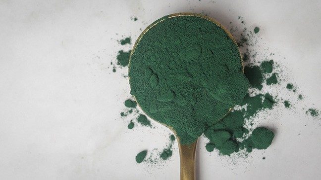 Gold spoon with dark green powder on a marble surface.
