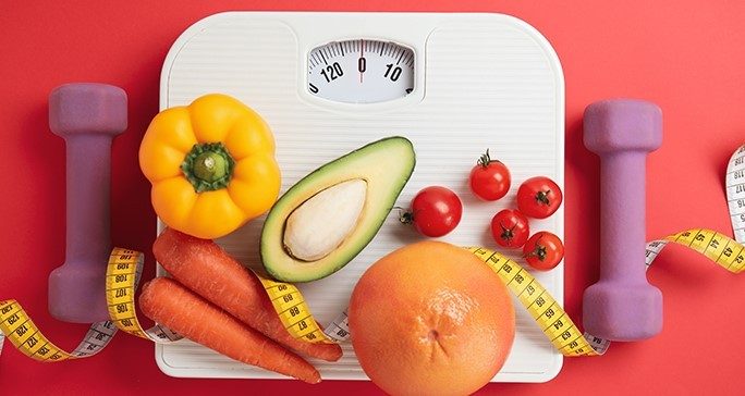 A white scale draped with a yellow measuring tape sits on a red surface. The scale holds two carrots, half on an avocado, a yellow bell pepper, an orange, and five cherry tomatoes. Purple hand weights are on either side of the scale.