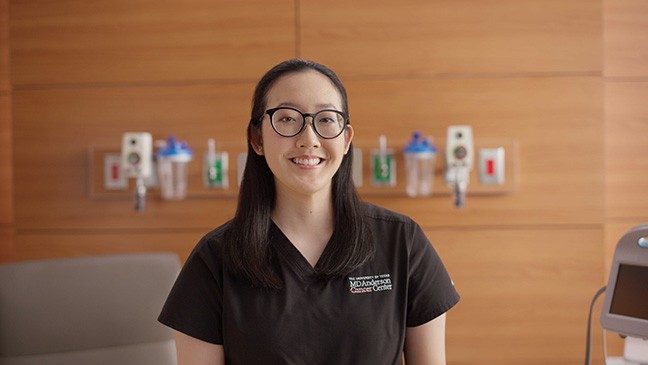 Smiling woman with black hair and glasses wearing black MD Anderson scrubs stands in an examination room.