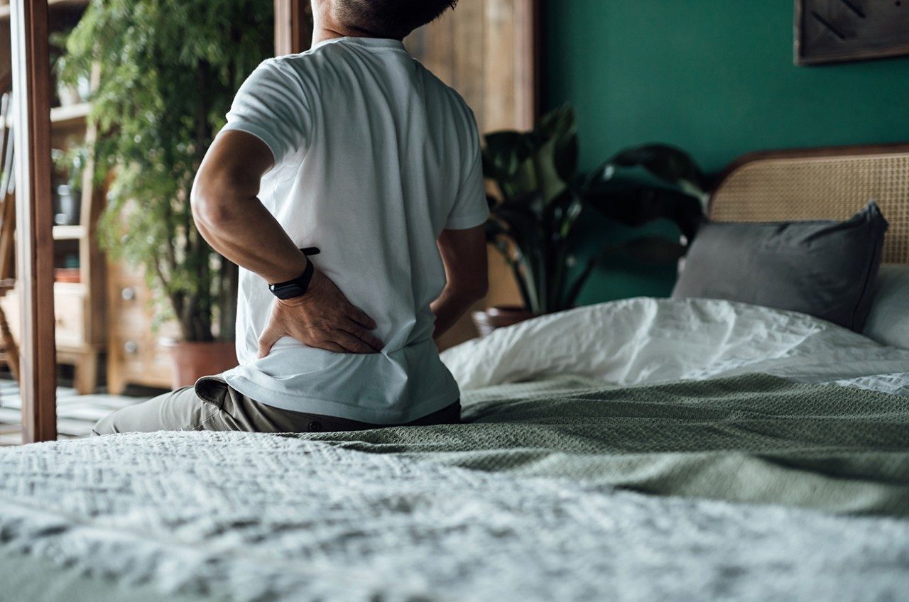 Unidentified man sitting on edge of bed holding his aching back