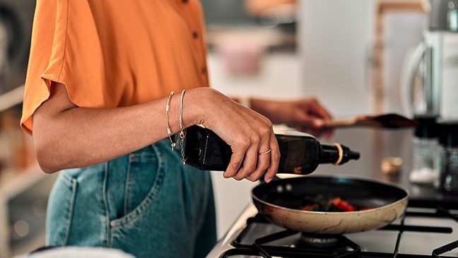 A person wearing an orange shirt and jeans pours oil into a skillet on a stovetop.