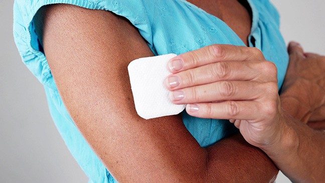 Person in light blue blouse applies a white nicotine patch to their upper arm.