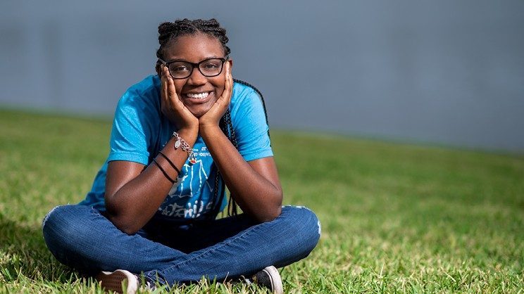 Akilah Lopez sits cross-legged in grass with lake in background