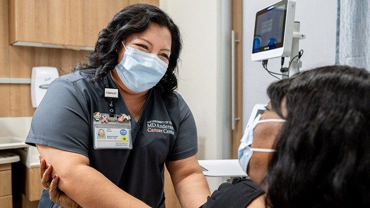 A woman wearing grey MD Anderson scrubs and a blue face mask works with a patient