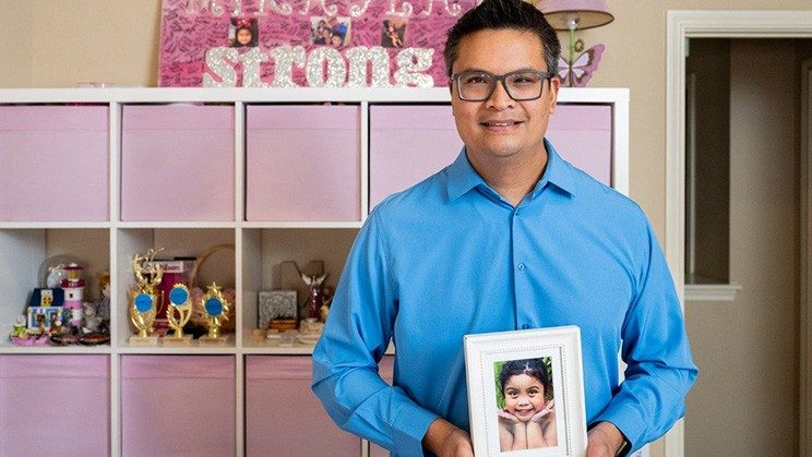 A man in a blue shirt stands in a room with pink furniture holding a framed picture of a smiling girl