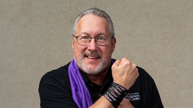 Doyle Bosque wearing a black MD Anderson embroidered polo and purple scarf, poses with arm across chest, displaying MD Anderson branded bracelets on his wrist.