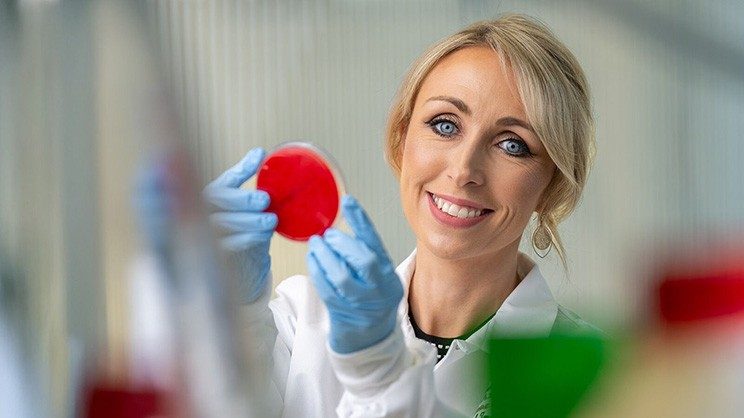 Smiling blonde woman in white lab coat and blue gloves holds a red petri dish.