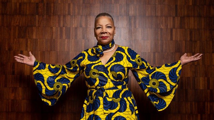 Marvetta Walker stands in a brightly-colored dress in front of a wooden textured wall