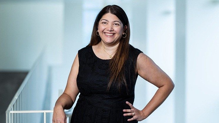 A smiling brunette woman in a black dress stands in a white hallway