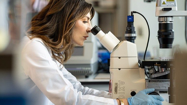 Woman with white lab coat and blue gloves looks into a microscope.