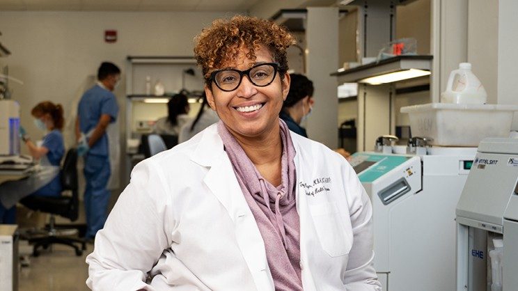 Toysha Mayer seated, wearing a white lab coat with lab equipment in background.