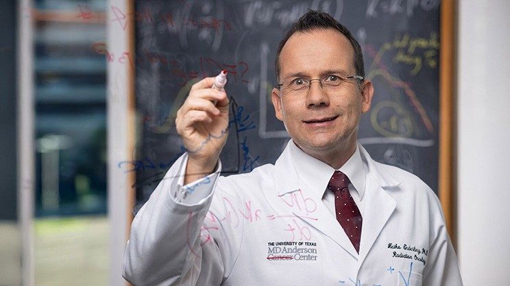 Man in white coat holds a marker and writes on a transparent board.
