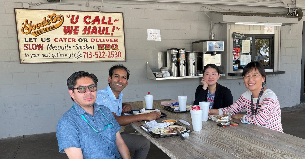 Four people sit at a wooden picnic table outside with a sign on the wall behind them that reads "Goode Co BBQ"