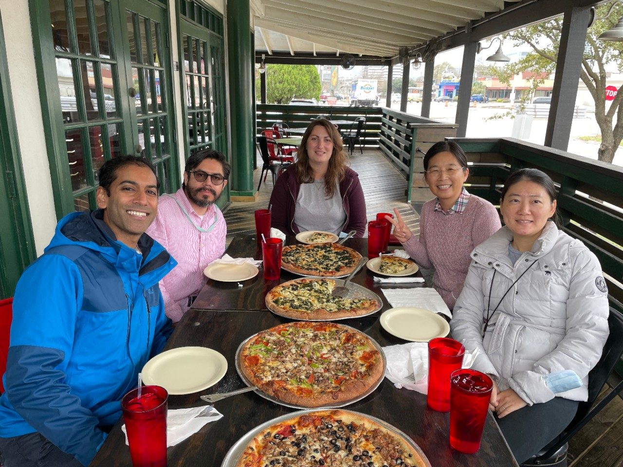 Five people sitting around a wooden picnic table on a restaurant porch with four pizzas on the table