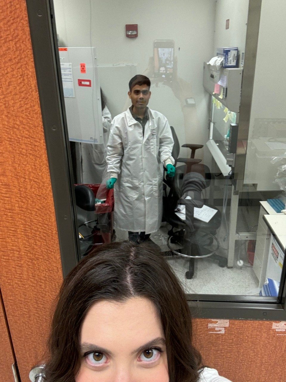 Selfie of one lab member with another lab member in the lab room behind them, seen through the door window