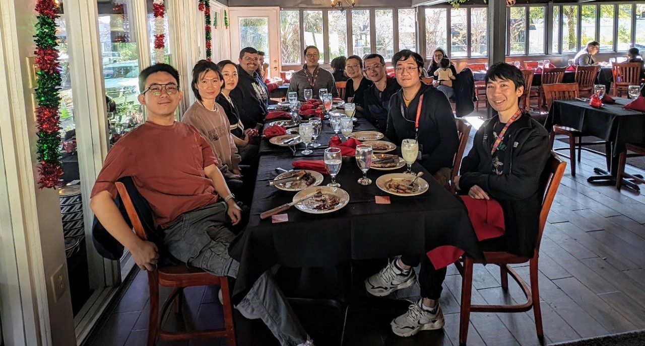 Nine people seated around a large rectangular table with a black tablecloth and red and green holiday decorations in the background