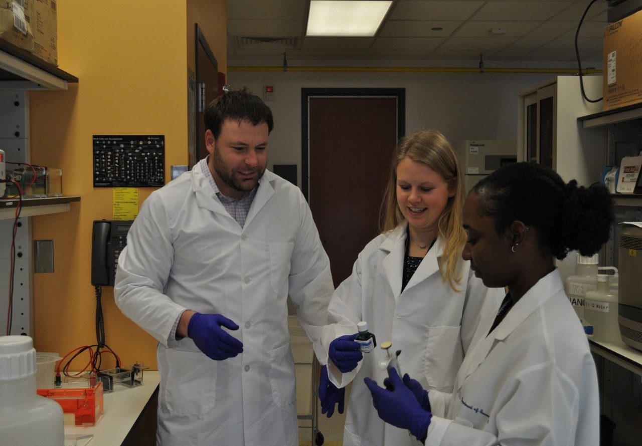 Three people in lab with white lab coats and dark blue-purple gloves