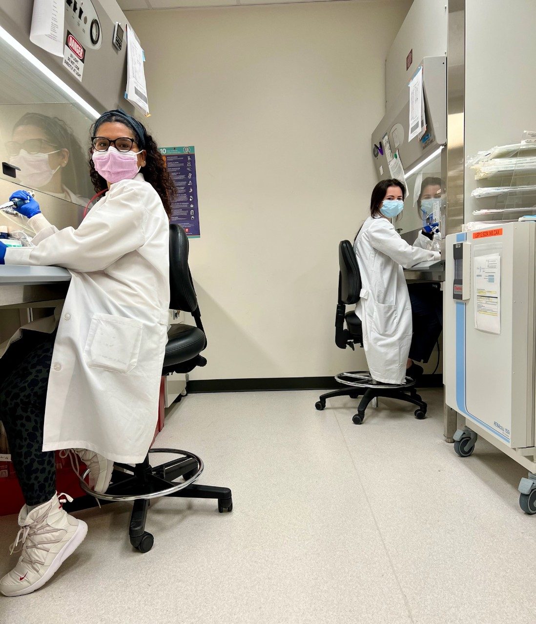 Two people with lab coats and masks working at hoods in a lab