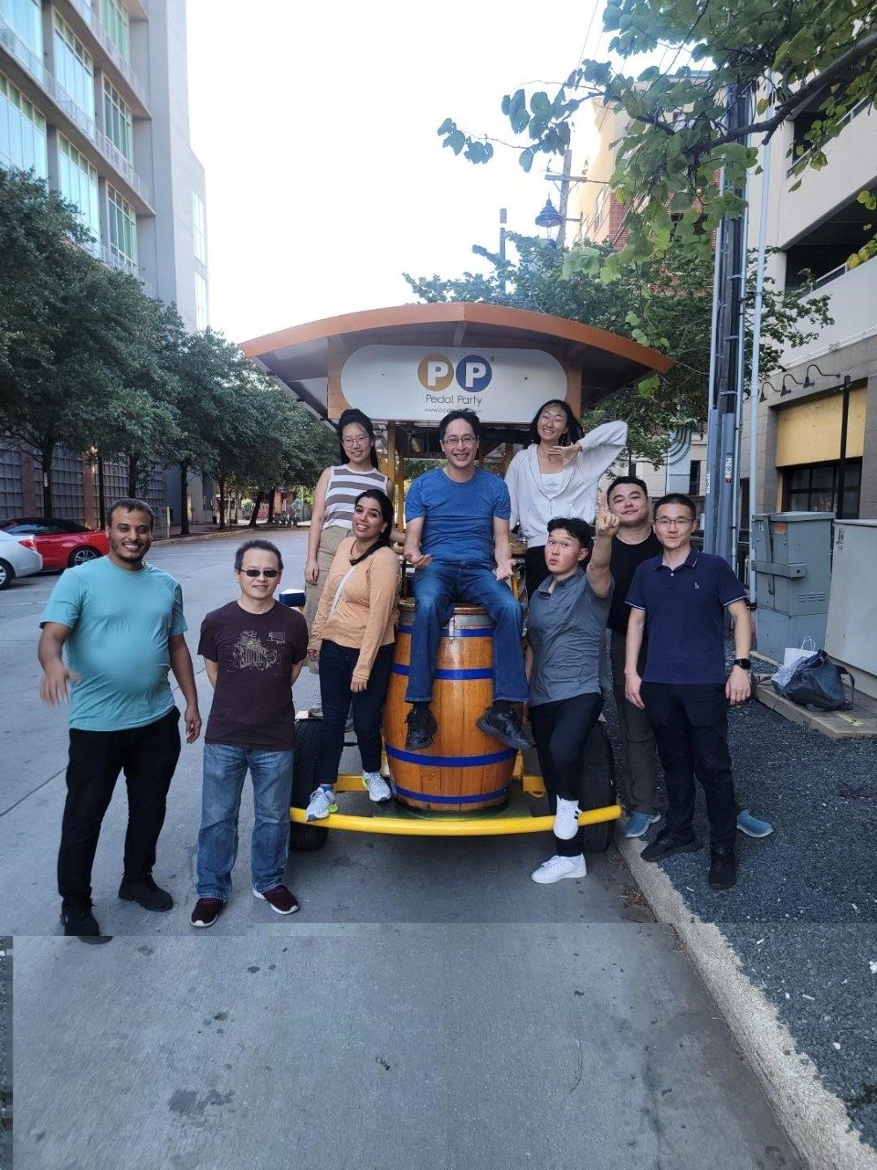 Group of people sitting on the end of a pedal party bus and standing around it, posed for a photo