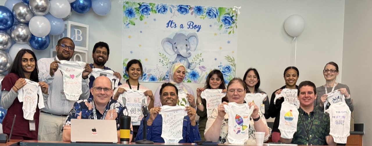 Lab members hold up decorated baby onesies. A sign on the wall behind them reads "It's a Boy"