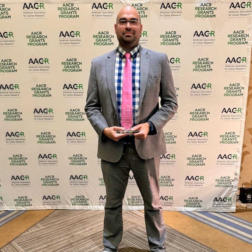 Dr. Reuben in front of an AACR branded wall in a grey suit with a plaid shirt and pink tie