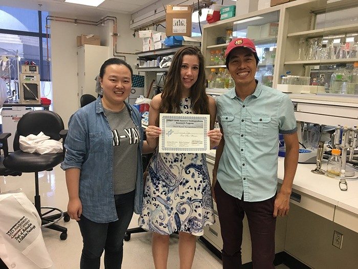 Three Tainer Laboratory members pose in the lab with the member in the middle holding a certificate